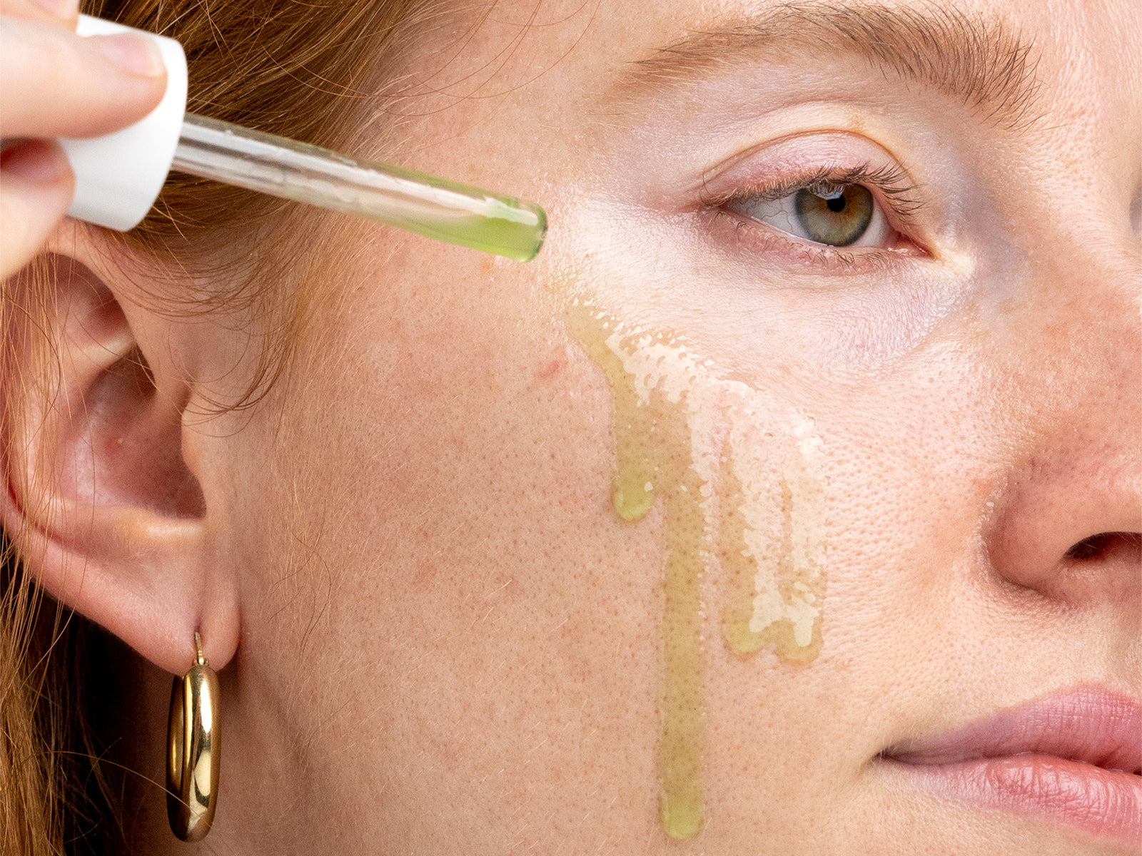 Nahaufnahme eines Gesichts, auf das mit einer Pipette ein grünes Serum auf die Wange geträufelt wird