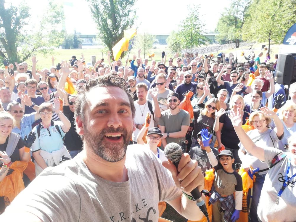 Ein Mann macht ein Selfie von sich und den Zuschauer:innen im Hintergrund und hält ein Mikro in der Hand.