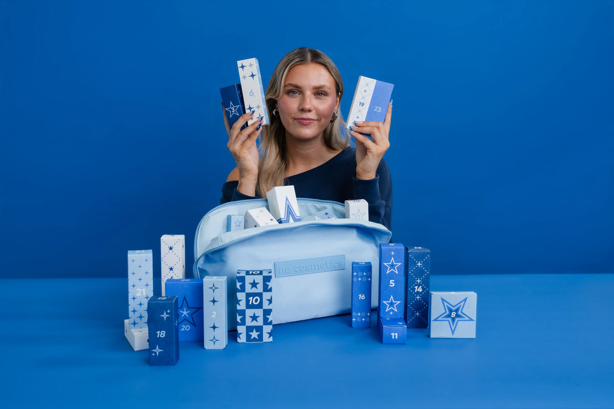 Frau präsentiert den Nø Cosmetics Adventskalender mit nummerierten Geschenkboxen und Nø Inside Magazin vor blauem Hintergrund.