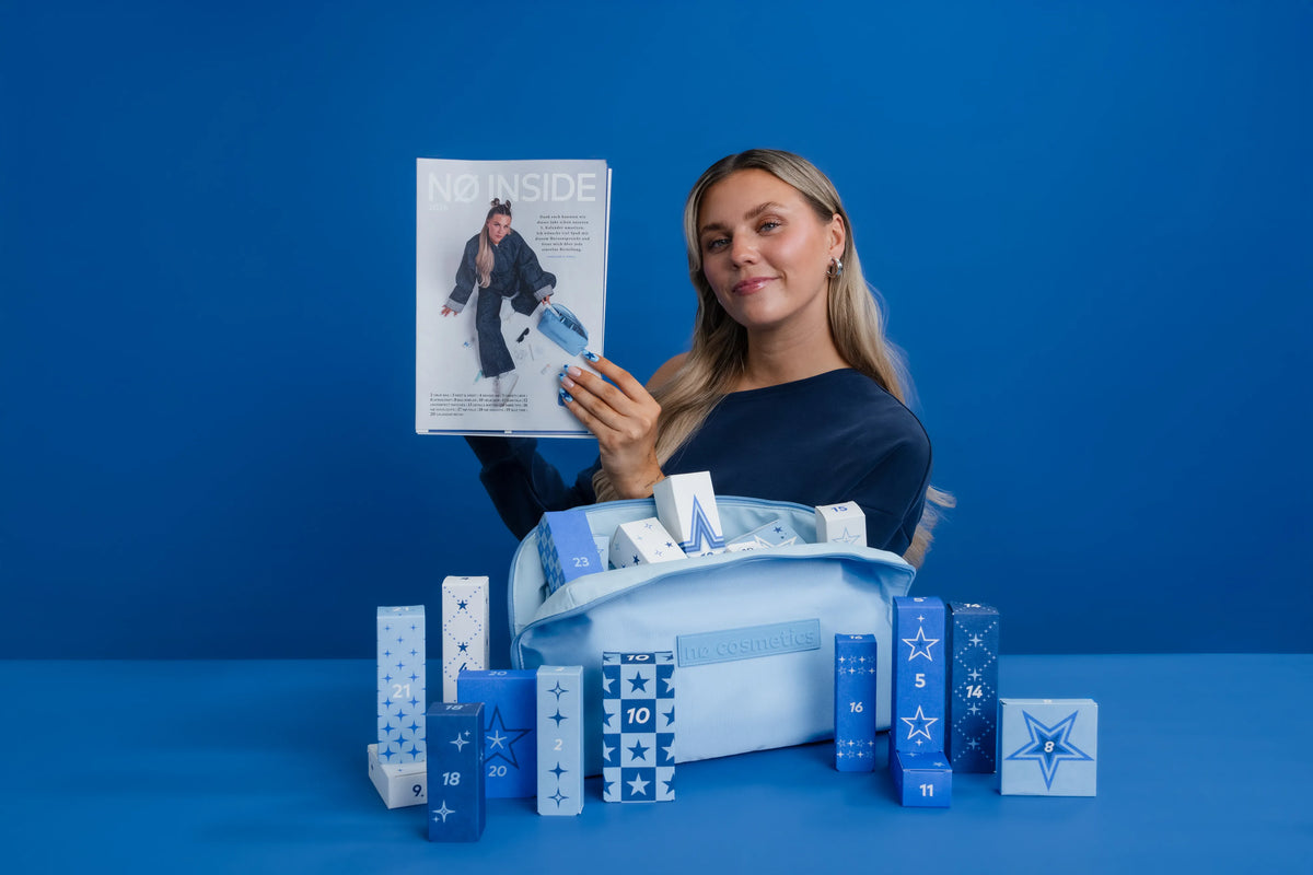 Frau präsentiert den Nø Cosmetics Adventskalender mit nummerierten Geschenkboxen und Nø Inside Magazin vor blauem Hintergrund.