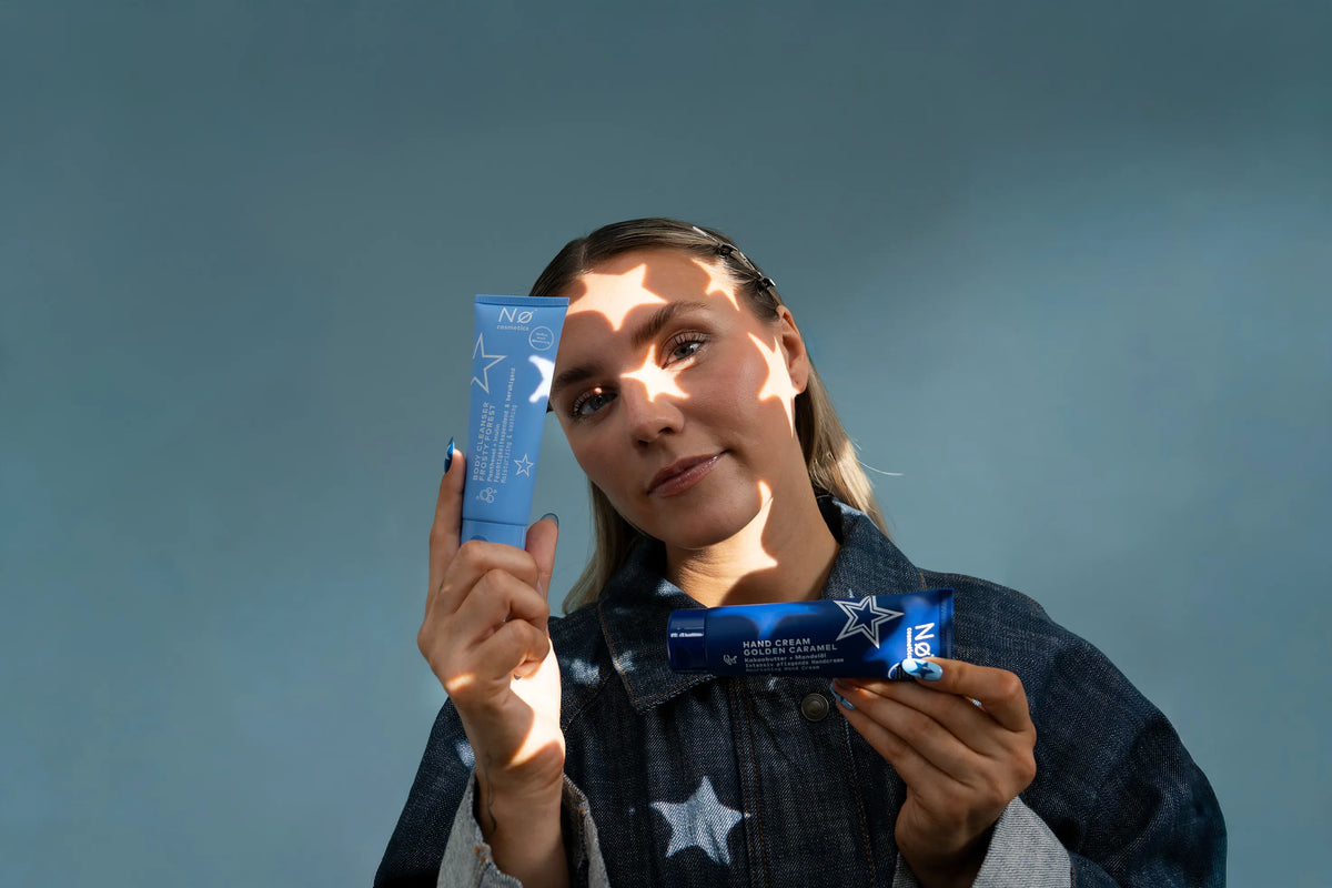 Frau hält zwei blaue Nø Cosmetics Hautpflegeprodukte vor hellblauem Hintergrund mit sternförmigen Lichtreflexen, horizontales Format.