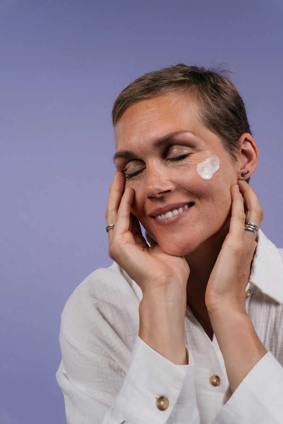 Lächelnde Frau mit aufgetragener Creme-Textur auf der Wange vor violettem Hintergrund.