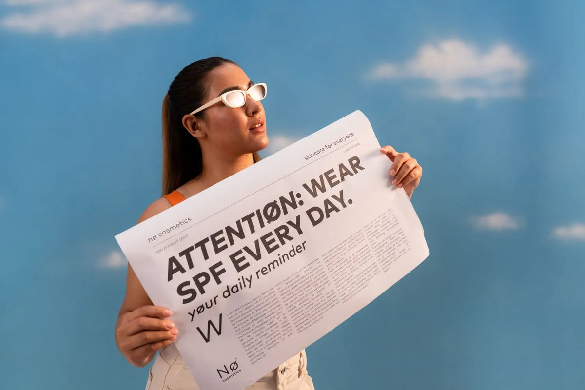 Frau mit weißer Sonnenbrille hält eine Zeitung mit der Aufschrift „ATTENTIØN: WEAR SPF EVERY DAY.“ vor blauem Himmel mit Wolken.